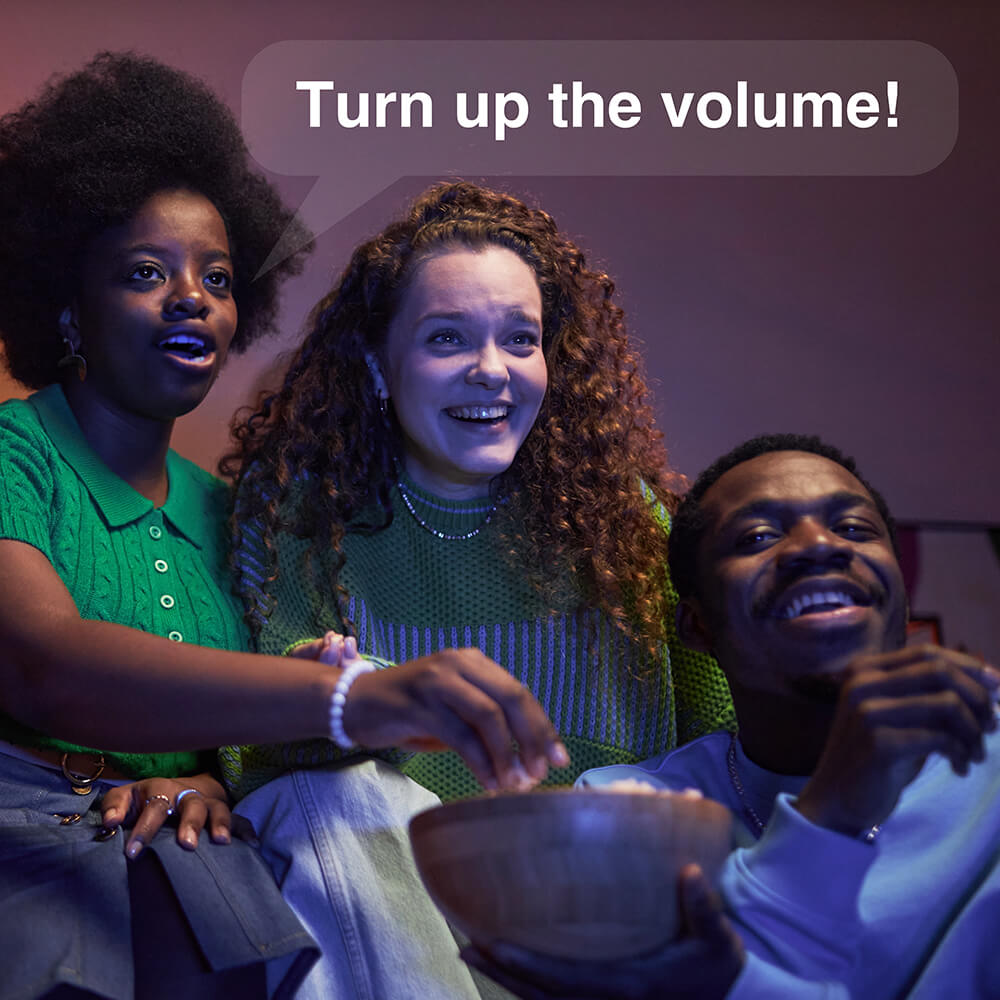Three friends using voice control to adjust TV volume while watching entertainment together on couch, showcasing smart TV voice command features